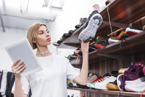 Blonde retailer holding digital tablet and shoe near shelves in second hand  - Foto, Bild