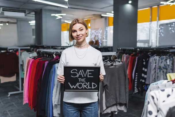 Smiling saleswoman holding board with save the world lettering in second hand  - Foto, afbeelding