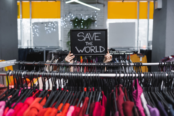 Oříznutý pohled na maloobchodníka držící desku s zachránit svět nápisy v blízkosti oblečení v druhé ruce  - Fotografie, Obrázek