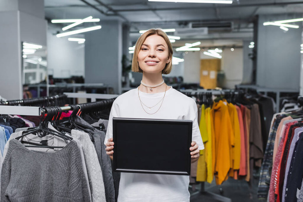 Positive saleswoman holding empty board near blurred clothes in vintage store  - Foto, immagini