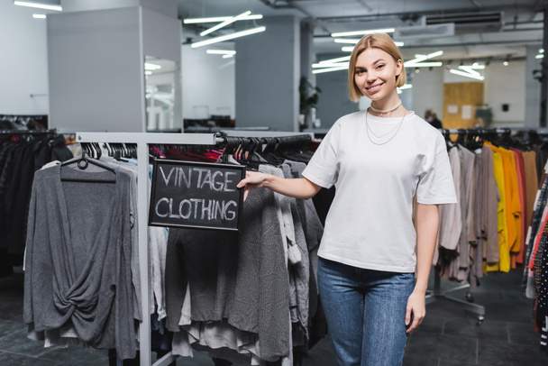 Smiling saleswoman holding board with vintage clothing lettering in retro store  - Photo, Image