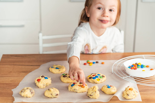 Little caucasian girl puts   multi-colored candies in shortbread cookies dough. Girl is smiling while making cookies at home - Photo, image