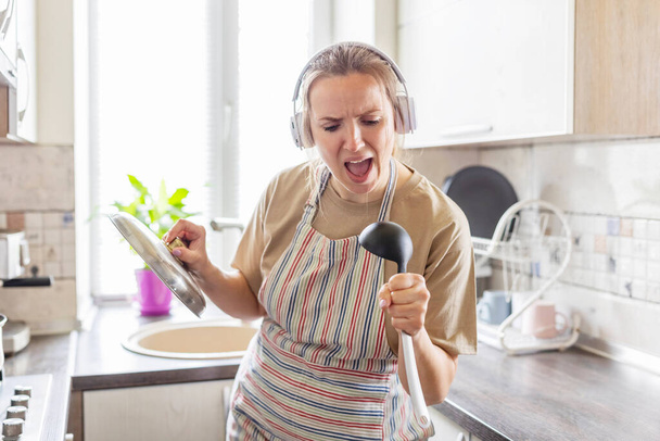 Hermosa mujer positiva en auriculares escuchando música y cantando en cucharón como micrófono mientras se cocina en la cocina. Relájese en la cocina. - Foto, Imagen
