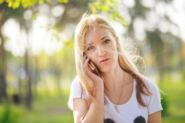 Blonde girl talking on the phone in the park in spring. Portrait of a beautiful fashionista mom with long hair talking to a friend on a smartphone. - Фото, зображення