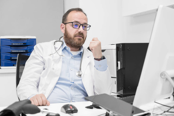 Portrait d'un thérapeute professionnel de la santé souriant assis sur le lieu de travail. Heureux médecin masculin confiant portant un manteau médical blanc et stéthoscope. - Photo, image