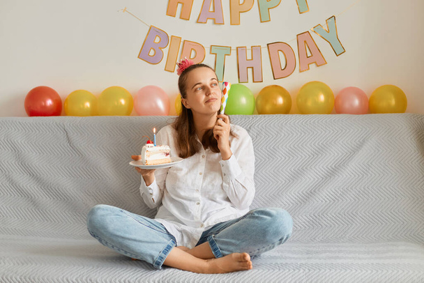 Image of pensive adorable young adult woman wearing white shirt celebrating birthday, sitting alone on cough with cake, holding party horn, looking away, dreaming. - Foto, afbeelding