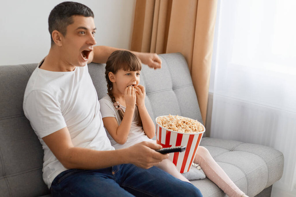 Portrait of young brunette guy with widely opened mouth watching horror movie at home with his daughter while sitting sitting on cough and eating popcorn in bucket. - Fotoğraf, Görsel