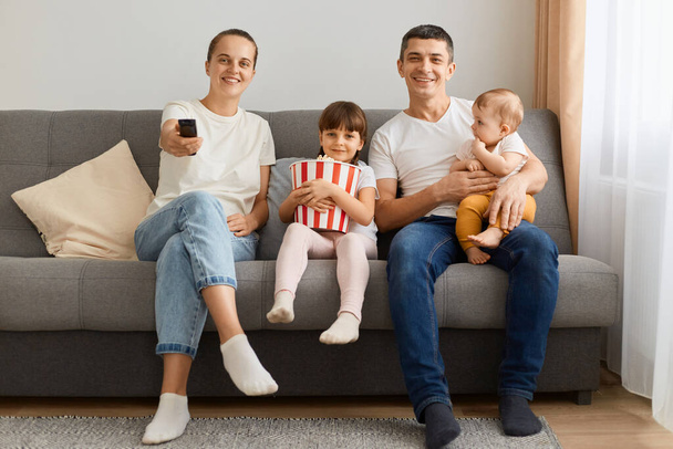 Indoor shot of man, woman and their daughter posing at home in living room and watching tv together, mother holding remote control, child with popcorn, enjoying time together. - Фото, зображення