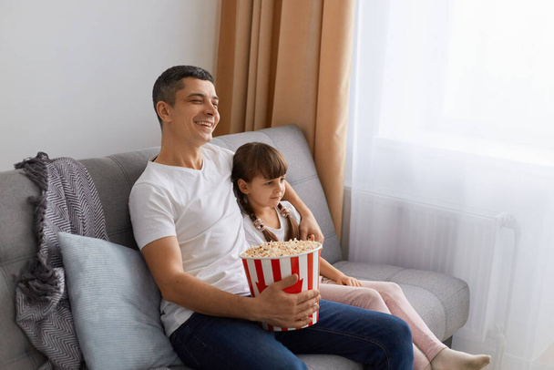 Side view portrait of satisfied positive brunette man wearing white t shirt sitting on sofa with his daughter and holding popcorn, family watching tv show or movie. - Foto, afbeelding