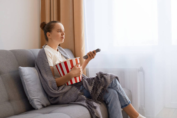 Side view portrait of beautiful young adult woman sitting on sofa in living room, watching a soap opera with sad upset facial expression, feeling sorrow. - Foto, Imagen