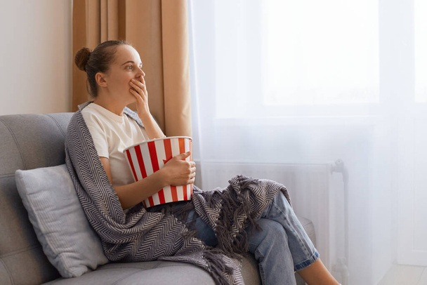Side view portrait of young adult woman spending time at home sitting on sofa in living room, watching television, scary or horror movie, in panic eating popcorn bowl. - Fotografie, Obrázek