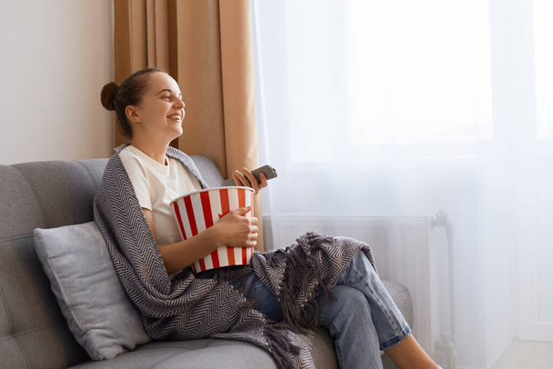 Horizontal shot of smiling happy positive woman sitting on sofa wrapped in blanket and watching tv, funny comedy show, expressing happiness, enjoying time spending at home alone. - Zdjęcie, obraz