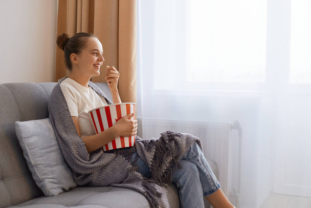 Side view photo of charming woman sitting on sofa and eating popcorn bowls, watching movie or interesting tv show, enjoying tasty snack while watch interesting tv show. - Foto, afbeelding