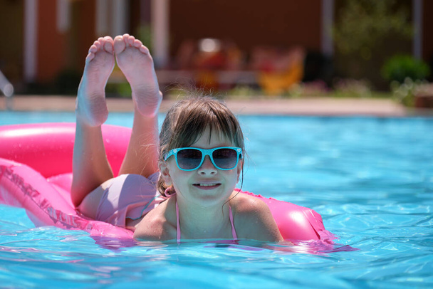 Happy child girl relaxing on inflatable air mattress in swimming pool on sunny summer day during tropical vacations. Summertime activities concept - Photo, Image