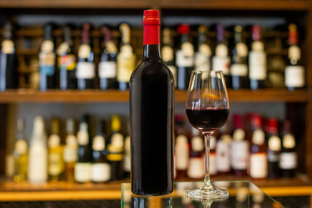Fles en glas rode wijn in een winkel met een verscheidenheid aan flessen op de achtergrond uit focus - Foto, afbeelding