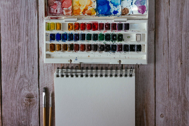 Palette of watercolor paints, blank paper and brushes on wooden background.  Artist's work space. Copy space. Selective focus. - Фото, изображение
