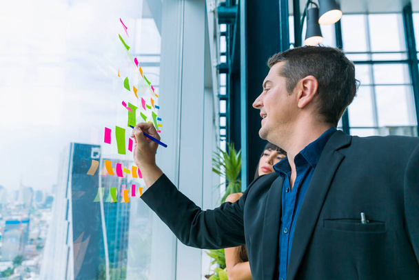 Geschäftsleute, die arbeiten und Post-it-Notizen verwenden, um Ideen am Glasfenster im Büro auszutauschen. Unternehmer und Unternehmerin arbeiten im Projektplanungsausschuss. - Foto, Bild