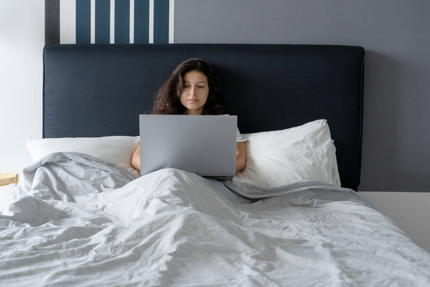 Beautiful brunette girl in bed with a laptop. Work from morning to late night. Electronic gadgets before bedtime or in the morning - Zdjęcie, obraz