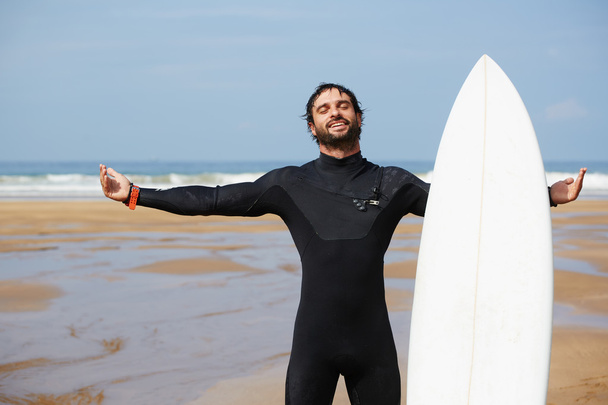 Young surfer feeling so happy, attractive surfer enjoying perfect sunny day standing on the beach and holding with one hand his surfboard, feeling good and happy with healthy lifestyle, filtered image - Foto, Imagen