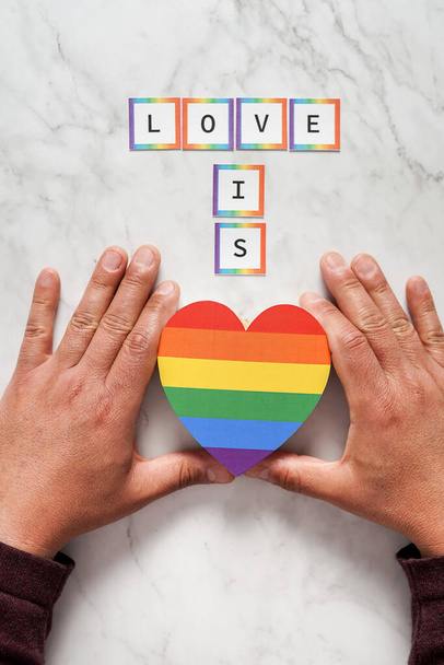 Hands of a mature man placing a heart with the colors of the LGBTQ movement under the phrase Love is. Concept of tolerance, inclusion and diversity - Fotografie, Obrázek