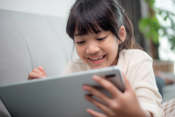 インターネット上でタブレットゲームをプレイしている小さな女の子、子供はソファに座ってオンラインで友人と見たり話したり、朝のリビングルームでリラックスしたり、新しい技術の概念を持つ子供 - 写真・画像