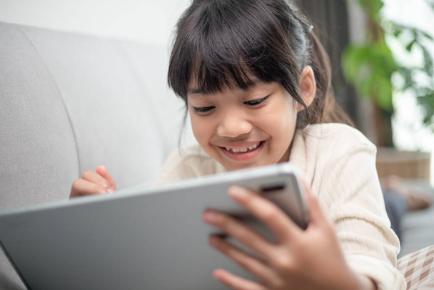 インターネット上でタブレットゲームをプレイしている小さな女の子、子供はソファに座ってオンラインで友人と見たり話したり、朝のリビングルームでリラックスしたり、新しい技術の概念を持つ子供 - 写真・画像