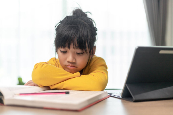 小さなアジアの女の子一人で座って退屈そうな顔で外を見ると、就学前の子供は宿題で悲しい退屈とテーブルの上に頭を置く、甘やかされた子供 - 写真・画像