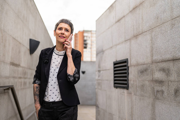 One woman modern mature caucasian female businesswoman with short hair using mobile phone making a call talk while walking on the stairs by building in day against concrete or stone wall copy space - Foto, afbeelding