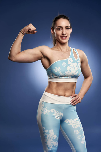 Young athletic woman posing on blue background, in gym attire - Φωτογραφία, εικόνα