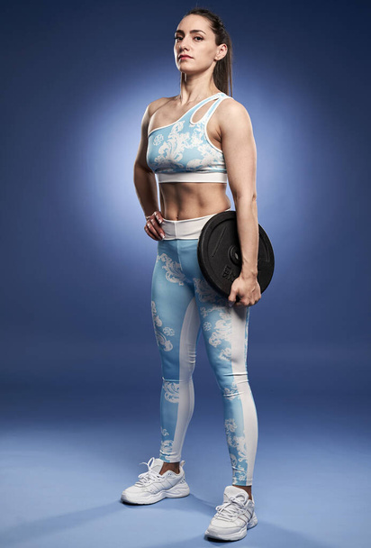 Strong woman with a gym plate posing full length on blue background - Fotó, kép
