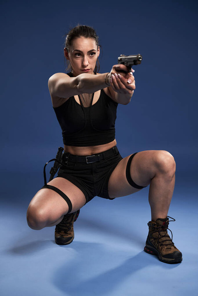 Athletic adventurer young woman in black shorts and top, with a gun and tactical knife, on blue background - Fotó, kép