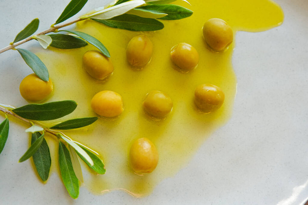 Оливки та оливкова олія. Зелені оливки та оливкова гілка в оливковій олії в керамічній сірій тарілці крупним планом. Холодна пресована натуральна олія
 - Фото, зображення