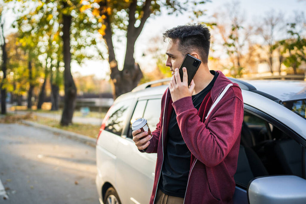 Giovane uomo caucasico in piedi vicino alla sua auto in autunno giorno o sera in possesso di una tazza di caffè da asporto guardando di lato durante l'attesa per strada in città copiare spazio facendo una telefonata parlando - Foto, immagini