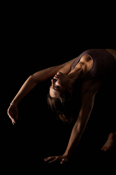Fit woman practicing yoga poses. Side lit half silhouette girl doing exercise in studio against black background. - Zdjęcie, obraz