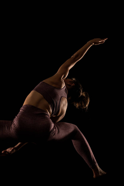 Fit woman practicing yoga poses. Side lit half silhouette girl doing exercise in studio against black background. - Foto, imagen