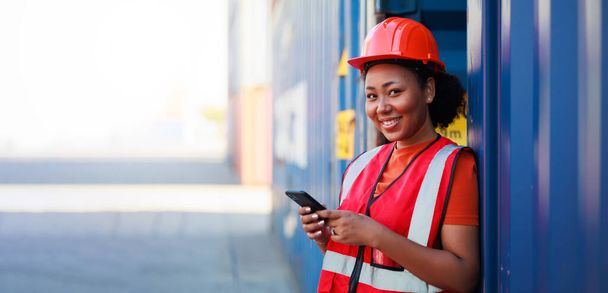 Online working by smartphone. Black female dock worker control loading containers box from cargo at warehouse container yard. Marine and carrier insurance concept. logistic shipping yard - Fotó, kép