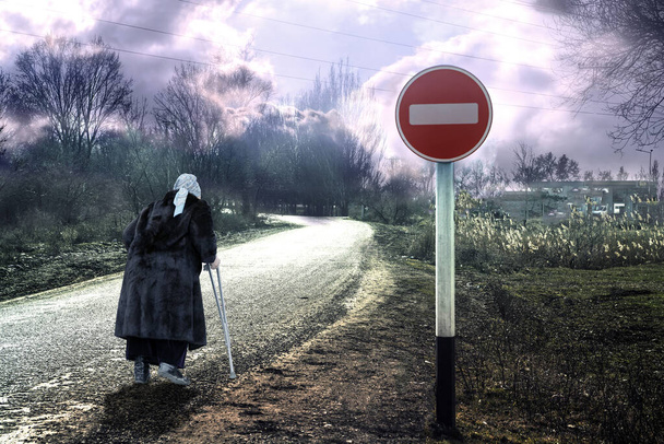 Une femme âgée avec une béquille sur la route sous un panneau interdisant. - Photo, image