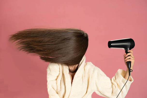 Woman drying her long hair with electric fan on a pink background. - Фото, изображение