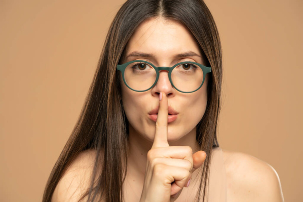 Портрет молодої жінки пальцем, що закриває рот губ, не каже, щоб камера ізольована на бежевому кольоровому фоні
. - Фото, зображення