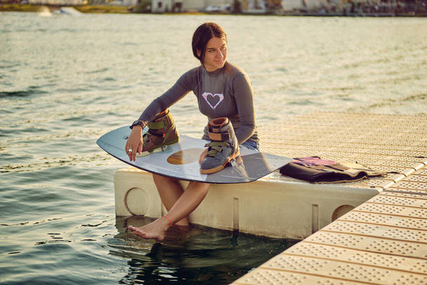 Brunette girl in a gray turtleneck and blue denim shorts is posing with her wakeboard on a pier of the coastal zone. - Foto, afbeelding