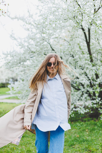 A young woman with blond hair enjoys a blooming spring garden. Travel, spring break. Fashionable style. A woman in sunglasses and a beige trench coat runs through a flowering park. Selection focus - Fotó, kép