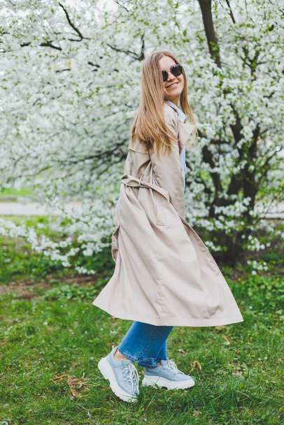 A young woman with blond hair enjoys a blooming spring garden. Travel, spring break. Fashionable style. A woman in sunglasses and a beige trench coat runs through a flowering park. Selection focus - 写真・画像