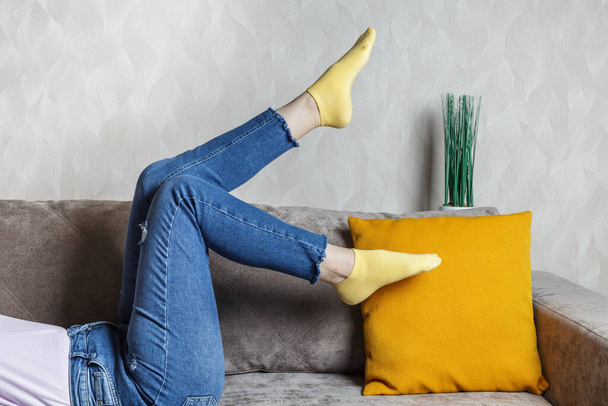 piernas delgadas de mujer en jeans y calcetines amarillos en el sofá. Sofá ligero y almohada naranja. - Foto, Imagen