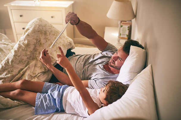Lets watch that next. Shot of a father and his little son using a digital tablet while lying in bed at home. - 写真・画像
