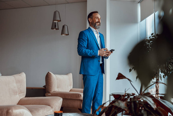 Mosolygó szakállas férfi okostelefonnal áll a modern irodahelyiségben - Fotó, kép