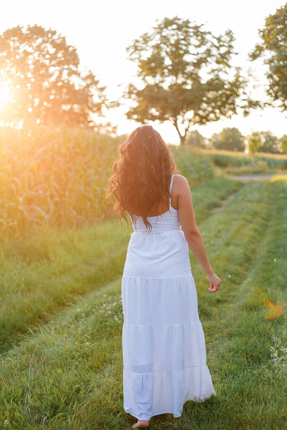 Portrait of a beautiful dark-haired curly young woman 30 years old in a white sundress in a summer field at sunset. Beautiful summer model. - Fotó, kép