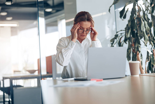 Moe zakenvrouw in bril met hoofdpijn werkend op laptop op haar werkplek op modern kantoor - Foto, afbeelding