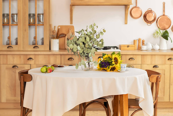 Sala de jantar elegante com mesa de madeira e cadeiras. Cozinha aconchegante decorada com decoração de verão e mesa que define flores e frutas de verão. Jantar de família de férias. interior da cozinha com utensílios, panelas - Foto, Imagem