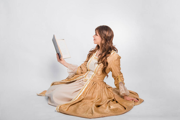 Full length portrait of a young woman in a puffy gold dress in the rococo era, posing while sitting with a book in her hands, isolated on a white background. The girl is reading a book. - Foto, Imagen
