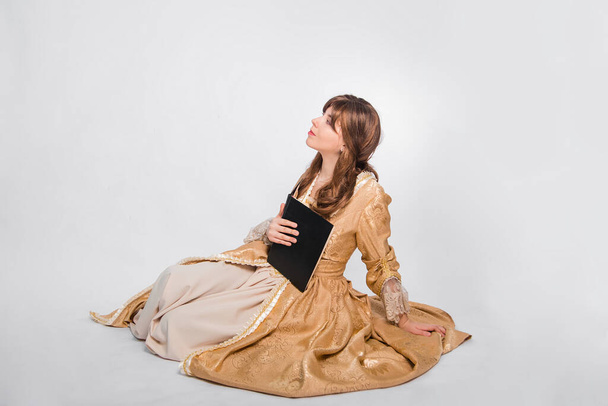 Full length portrait of a young woman in a puffy gold dress in the rococo era, posing while sitting with a book in her hands, isolated on a white background. The girl is reading a book. - Foto, afbeelding